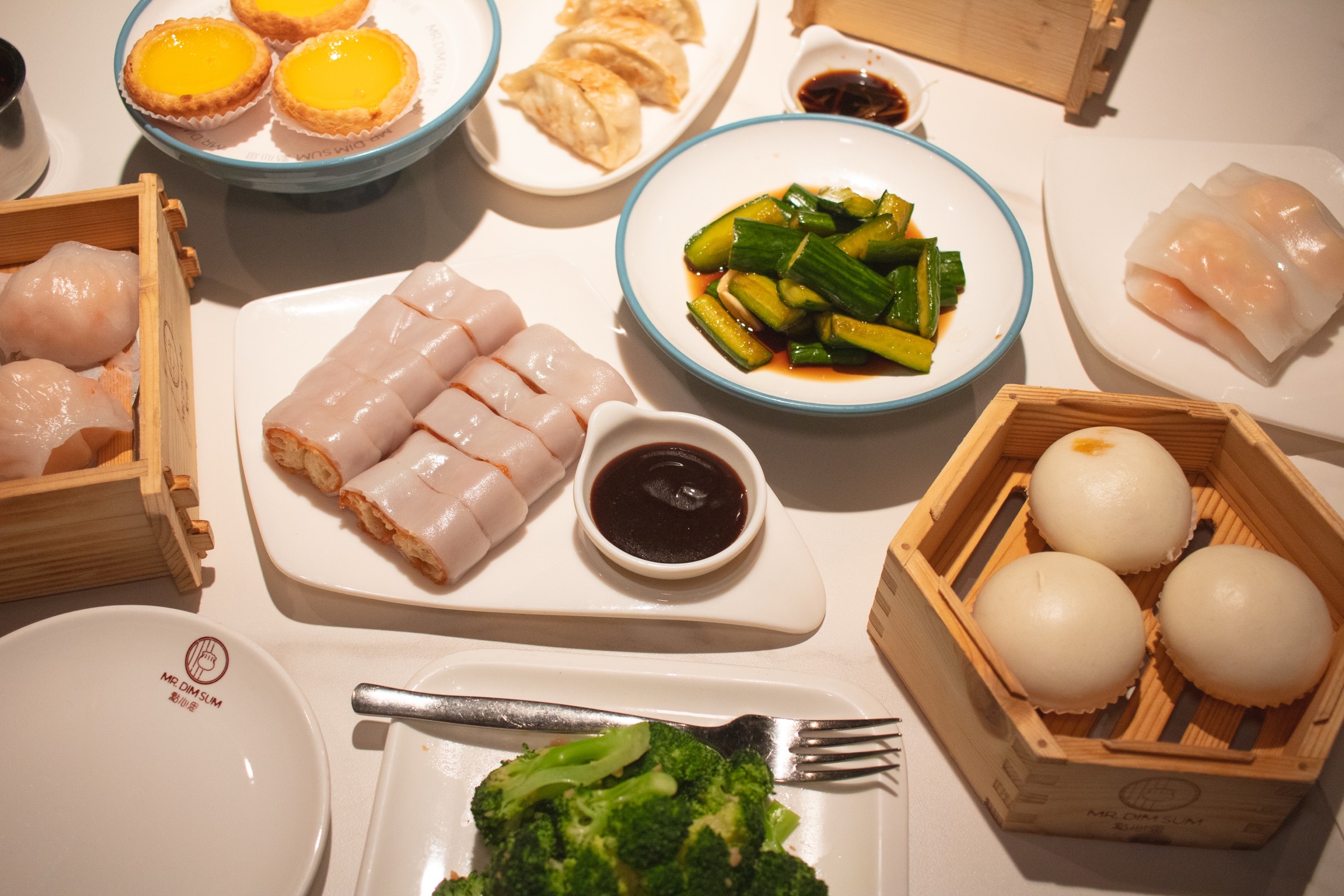 An overhead spread of Mr. Dim Sum dishes including har gow, rice rolls, steamed buns, and stir fried greens